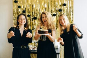 Drie mensen in zwarte kleding poseren voor een feestelijke achtergrond met gouden decoraties, terwijl één persoon een verjaardagstaart met het cijfer 